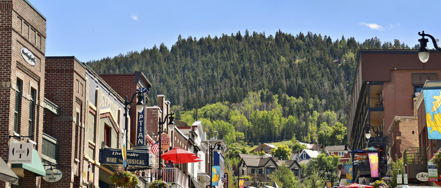 Street view of downtown Breckenridge, Colorado with brick buildings, shops, and forested hills in the background.