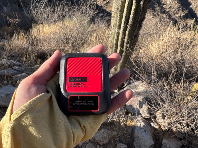  Close-up of a hand holding the Garmin inReach Messenger Plus in the desert, with cacti and dry vegetation in the background.