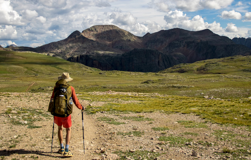 Backpacker wearing a large pack and using trekking poles hikes toward dramatic peaks in the San Juan Mountains.