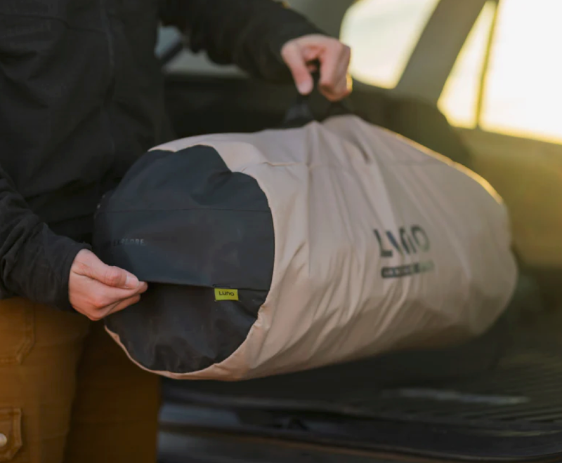 A person carrying a packed Luno mattress bag with a handle, preparing for a car camping trip at sunset.