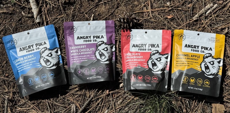  Four Angry Pika granola breakfast pouches—Lemon Berry, Cranberry White Chocolate, Chocolate Strawberry, and Caramel Apple—lined up on dirt and pine needles.