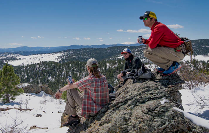 Three hikers sitting on rocks with Mountainsmith packs, enjoying snowy mountain views and drinks.