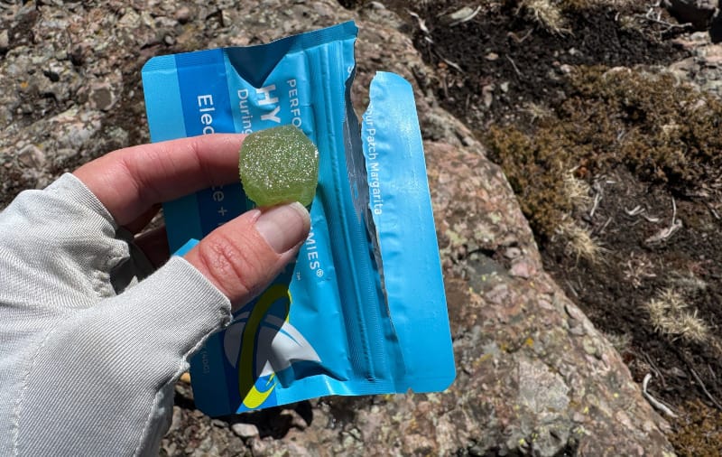  Close-up of hand holding a HydraFuel Hydration Gummy by Seattle Gummy Company above the product package on a rocky alpine trail.