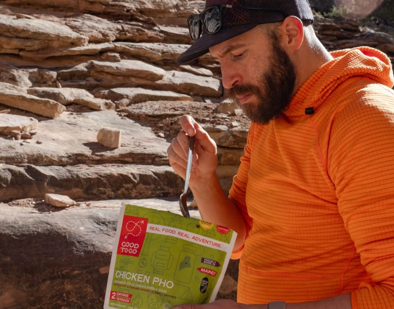 Backpacker in an orange hoodie eats Good To-Go Chicken Pho dehydrated meal beside red sandstone cliffs.