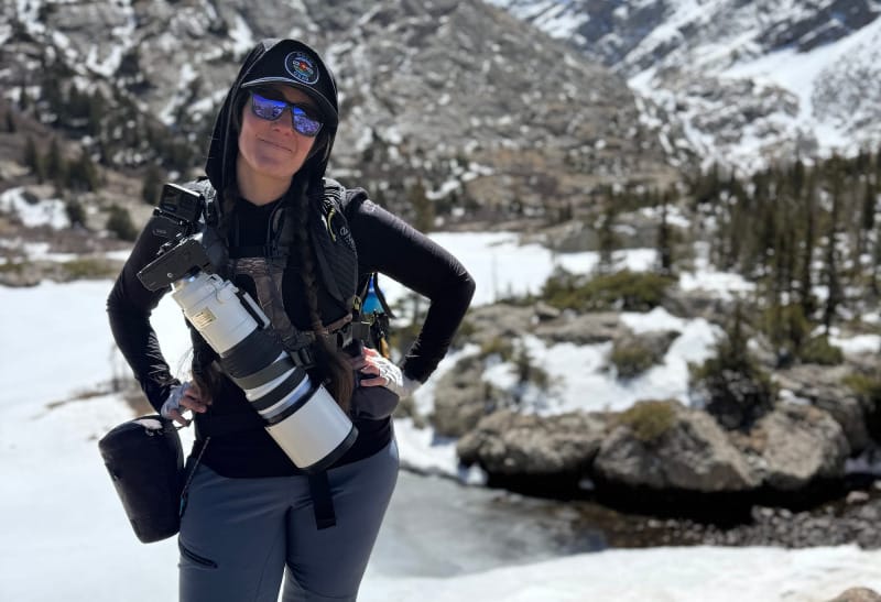 Photographer standing on a rock in the alpine with a large lens, camera, and Hyperlite Camera Pod mounted at the waist