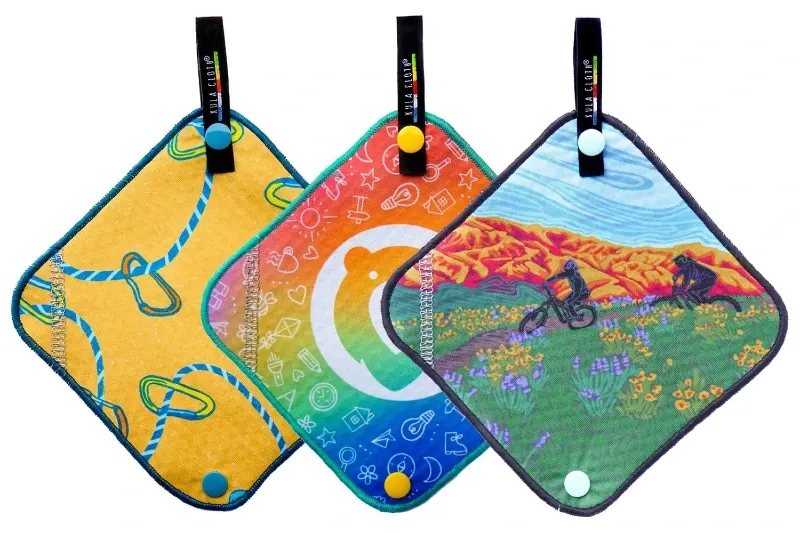 Three Kula Cloths hanging side by side, featuring a rope design, a rainbow design, and a mountain biking desert scene design.