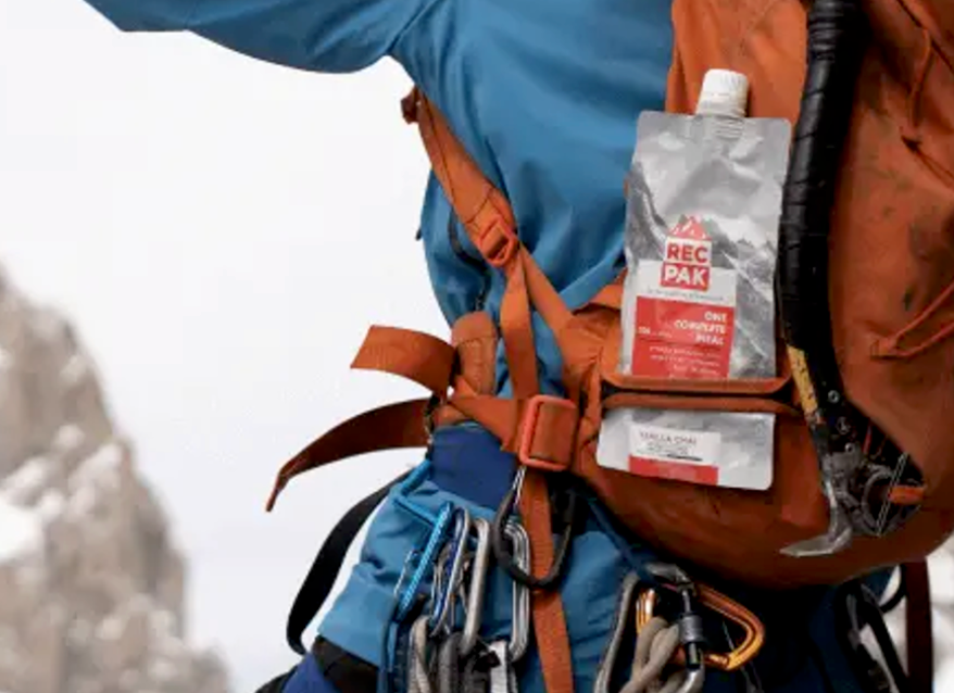 Close-up of a climber’s backpack with a RecPak meal pouch stored in the side pocket, surrounded by climbing gear and snowy mountain terrain.