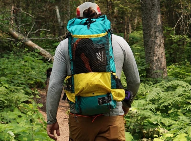 Back view of a hiker wearing a colorful Chicken Tramper Ultralight Gear backpack while walking through a green forest trail.