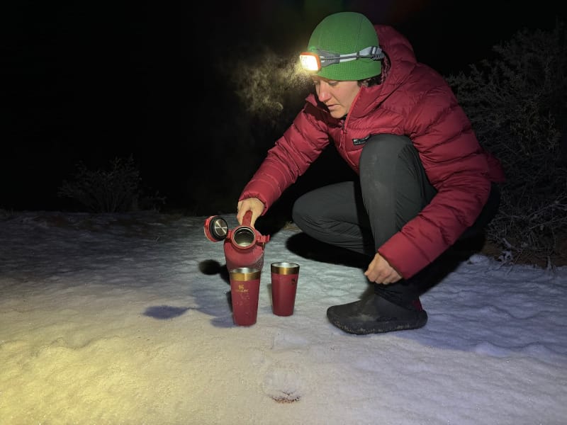 Backpacker wearing a red down jacket and headlamp pours hot drinks in the snow while camping at night.