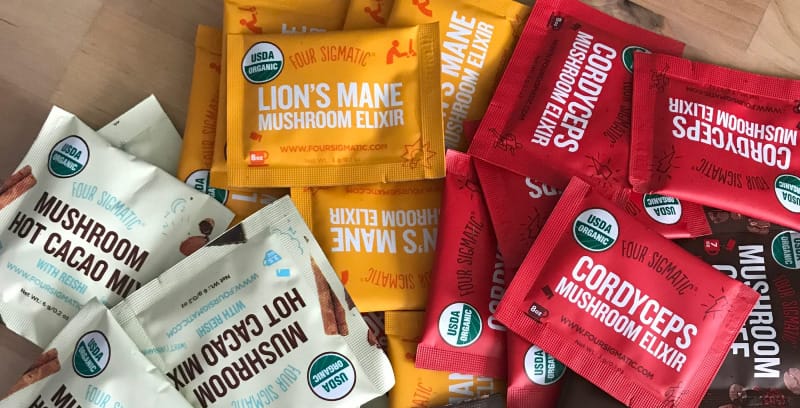 Assorted Four Sigmatic single-serve packets including Mushroom Coffee, Lion’s Mane Elixir, Cordyceps Elixir, and Mushroom Hot Cacao Mix spread on a wooden surface.