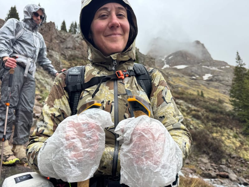 Smiling hiker wearing camo rain jacket and Adotec mitt stuff sacks on trekking poles in rainy mountain terrain