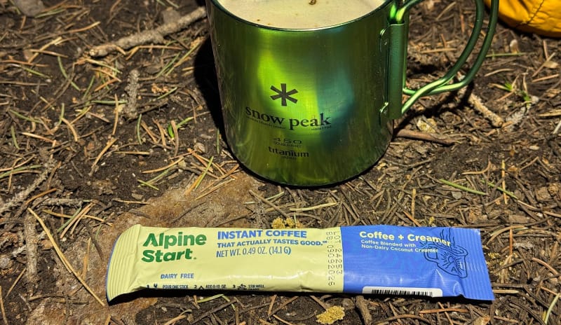  Alpine Start Coffee + Creamer instant packet on the ground next to a Snow Peak titanium mug filled with coffee at a forest campsite.