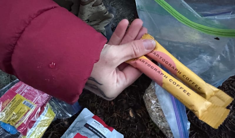  Hand holding two Cartograph Coffee instant espresso sticks next to backpacking food and resealable bags at a campsite.