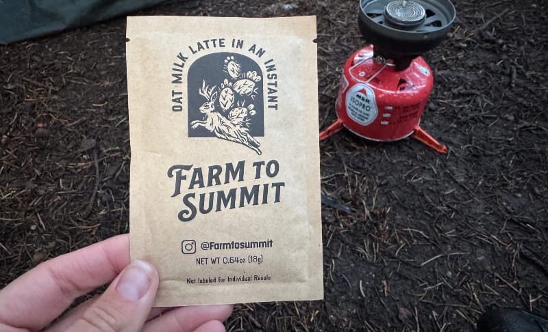  Farm to Summit instant oat milk latte packet held over forest ground with backpacking stove in the background.