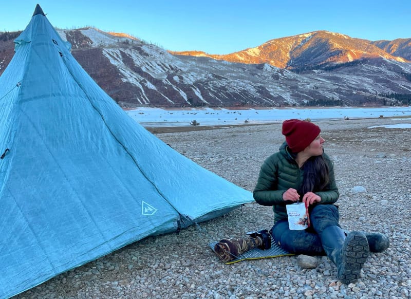  Camper sitting beside a pitched Hyperlite Mountain Gear UltaMid tent on a rocky riverbed with snowy mountains in the background, eating a backpacking meal.