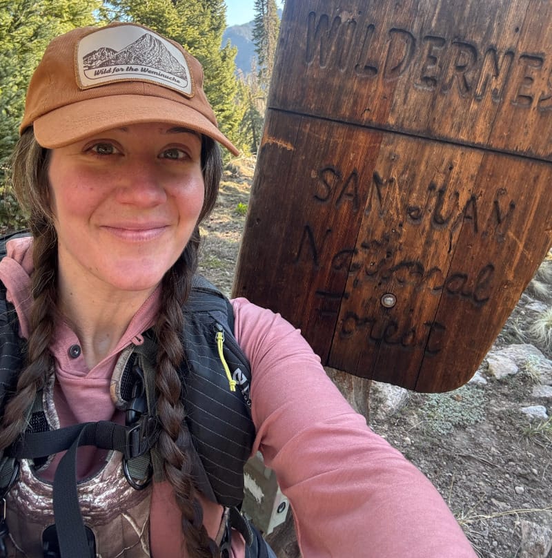 Hiker wearing the pink Ibex Springbok Hoodie in the Weminuche Wilderness next to a wooden trail sign in the San Juan Mountains.