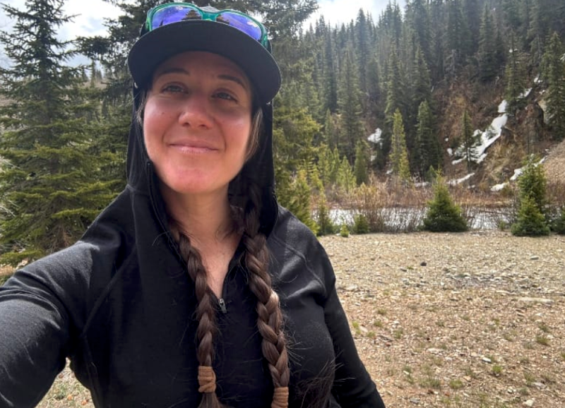 Person wearing a black Ibex Indie Hoodie with the hood up, standing in front of a high mountain valley with trees and snow patches.
