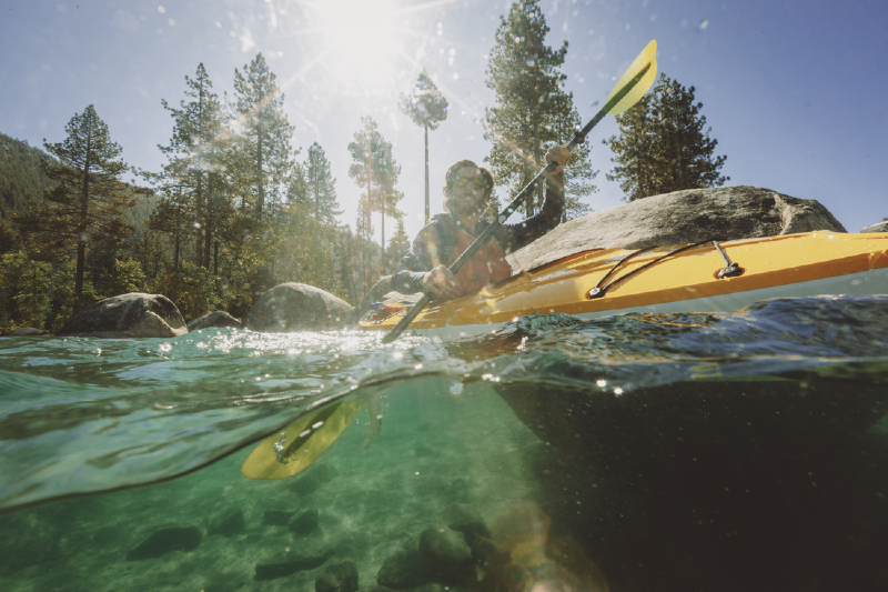 Person kayaking in crystal-clear mountain lake surrounded by pines and boulders, enjoying sunshine and outdoor adventure during REI’s 2025 Fourth of July Sale.