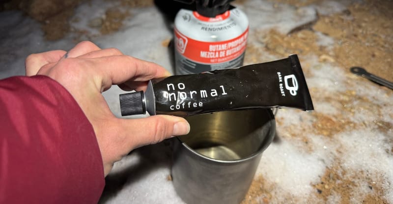 Hand holding a tube of No Normal Coffee dark roast in front of a backcountry camp stove with a metal mug on snow-covered ground.