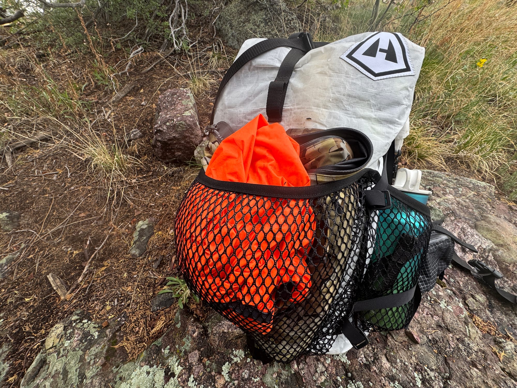 KUIU Chugach TR Rain Jacket and Pants packed in the mesh pocket of a backpack on a rocky trail.