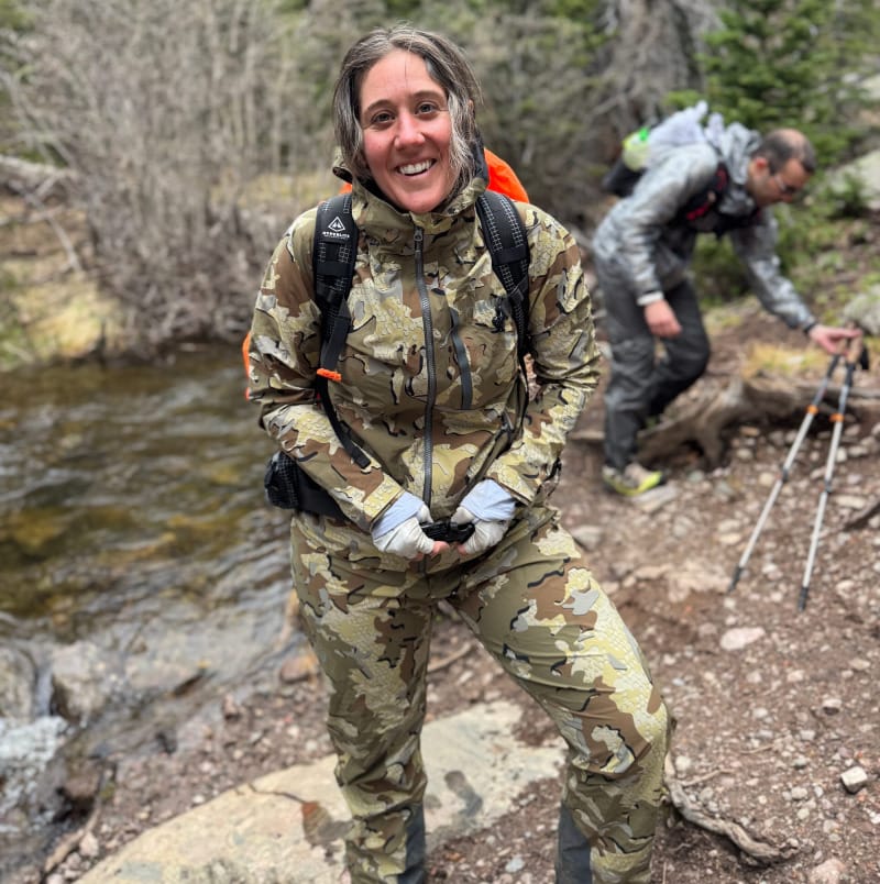 Hiker wearing the full KUIU Chugach TR Rain Jacket and Pants set near a creek in a high alpine forest.