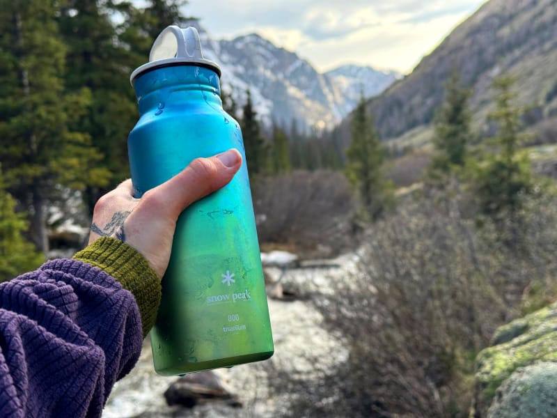 Hand holding the Snow Peak Aurora Titanium Water Bottle with alpine mountain scenery in the background