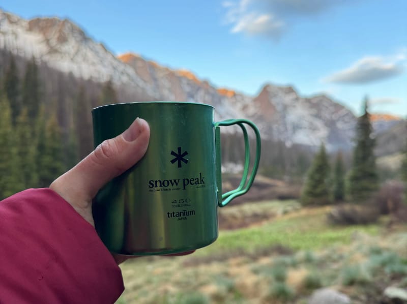 a backpackers holds the green ti-double titanium mug by snow peak in front of mountains during a camp.