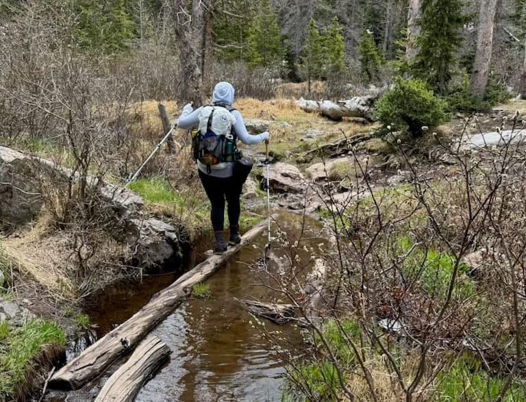 Hiker crossing a log bridge over a creek in the forest, wearing a pack and using trekking poles, with La Sportiva Spire boots visible.