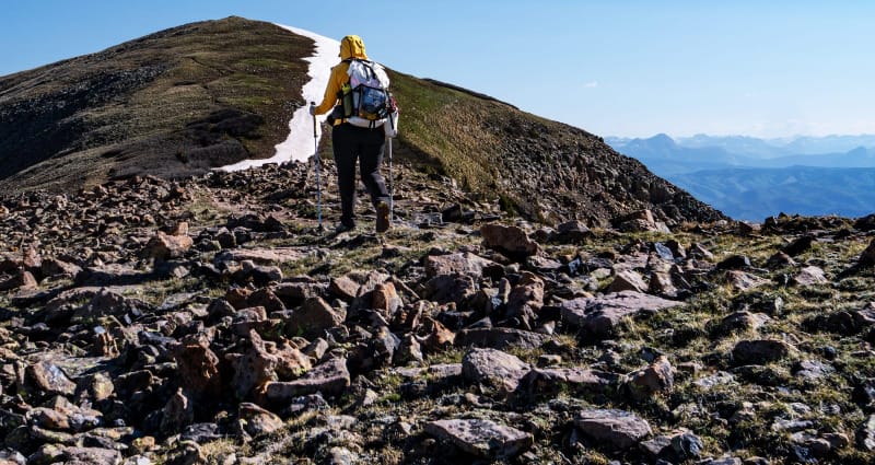  Back view of hiker walking across rocky alpine terrain toward a snow-covered summit wearing La Sportiva boots and a large backpack.