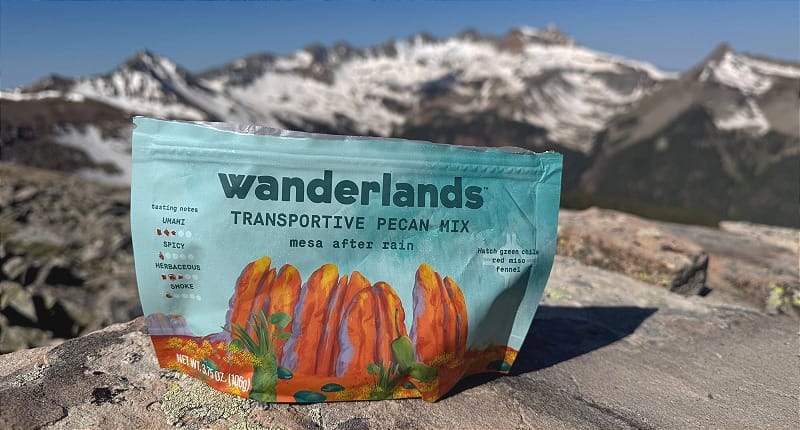  Wanderlands Transporative Pecan Mix “Mesa After Rain” flavor resting on alpine rock with snow-capped mountains in the distance under a clear sky.
