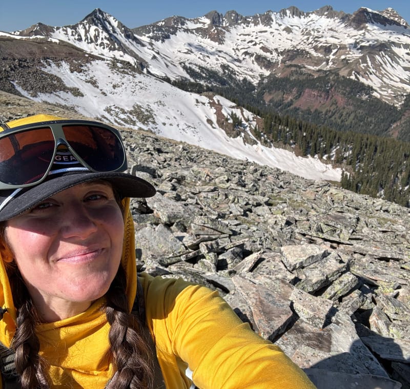Smiling hiker on a rocky summit wearing Vuarnet Altitude 01 sunglasses with snow-covered peaks behind.