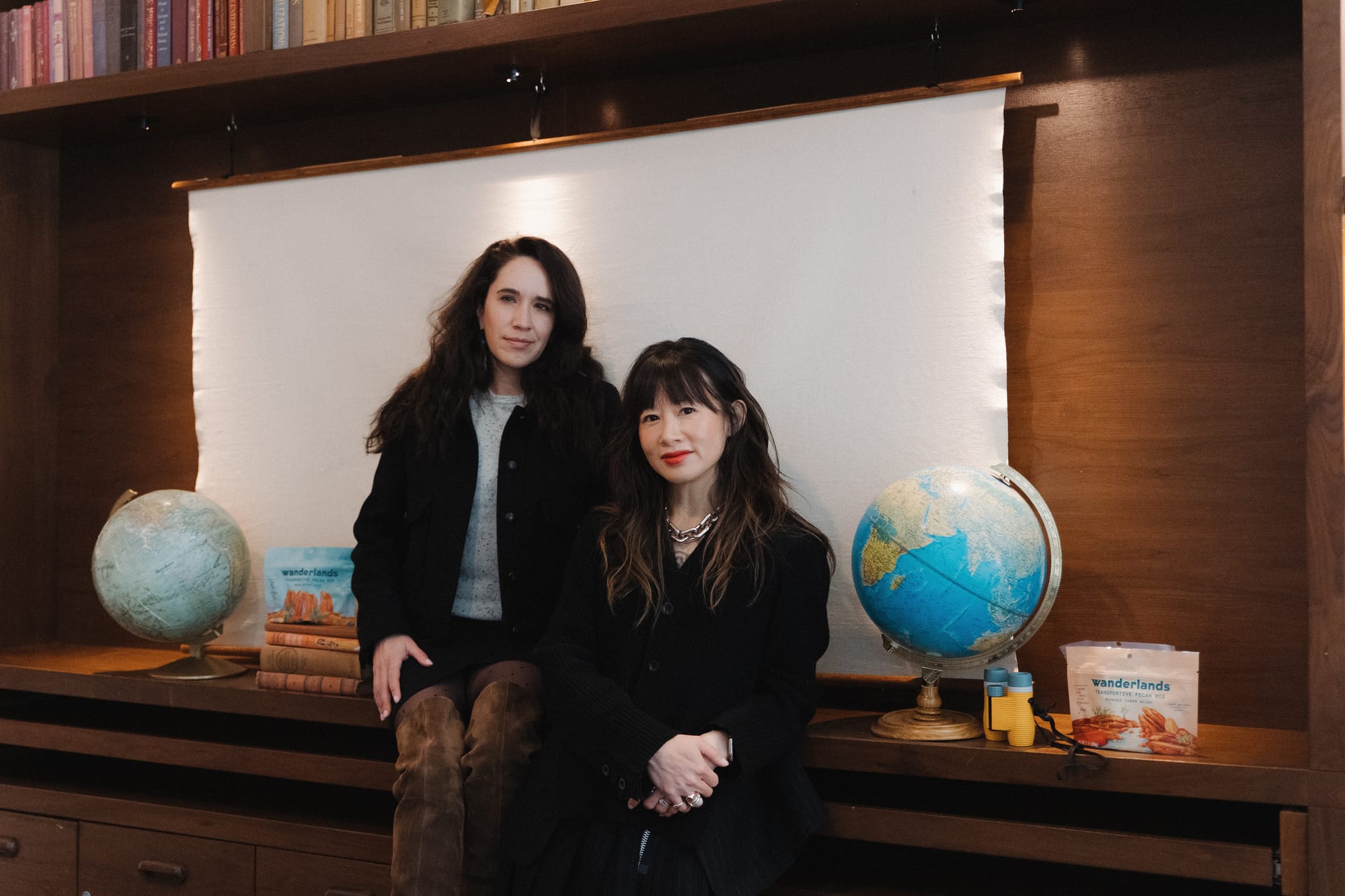  Portrait of Wanderlands founders Jane and Sofia seated in front of globes and Wanderlands pecan mix packaging, surrounded by books in a cozy, wood-accented room.