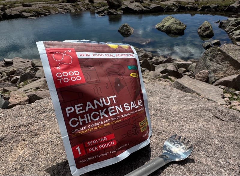 Good To-Go Peanut Chicken Salad meal pouch placed on a rock by a lake in the mountains with a spork beside it.