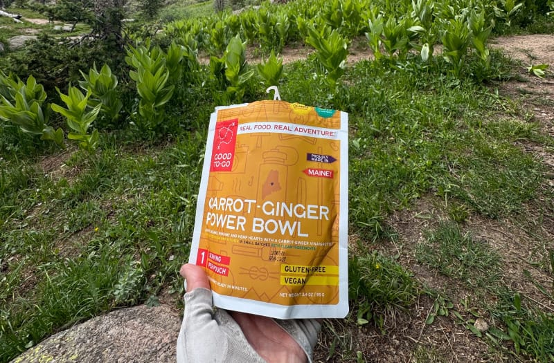 Backpacker holding a Good To-Go Carrot-Ginger Power Bowl meal pouch in a mountain meadow.