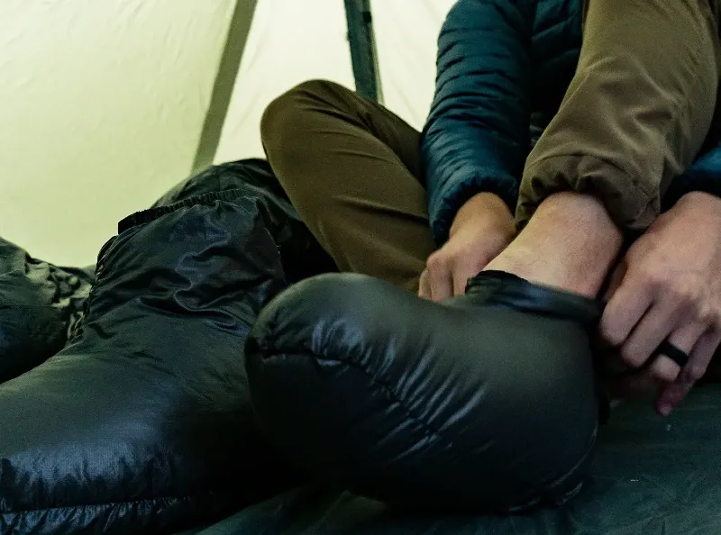 Person inside a tent putting on black down booties, dressed in insulated pants and jacket.