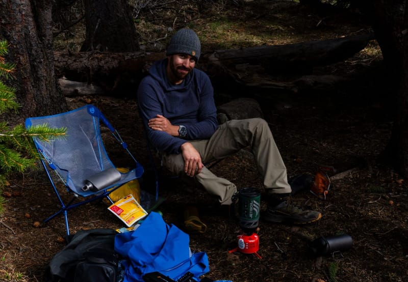 Backpacker relaxing at camp with a NEMO chair, cooking gear, and camera equipment scattered nearby.