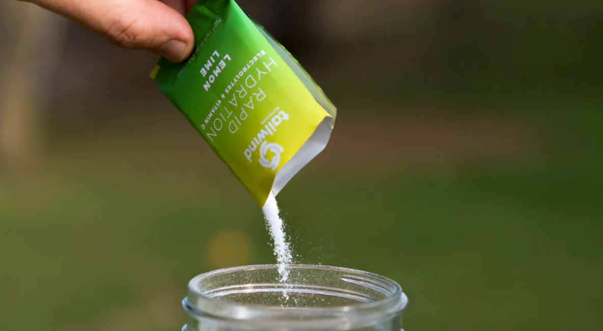 Hand pouring Tailwind Nutrition’s Lemon Lime Rapid Hydration powder into a clear water bottle with a green blurred background.