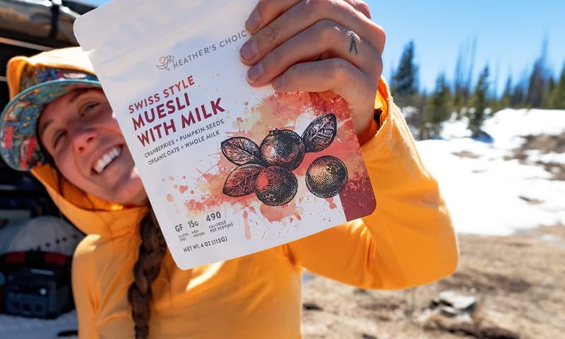 Backpacker smiling and holding up Heather’s Choice Swiss Style Muesli With Milk in front of their truck camper setup.