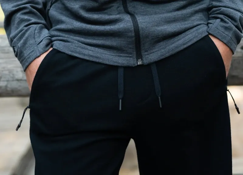 Close-up of a person wearing black merino wool joggers and a zip-up jacket, hands in pockets at a campsite.