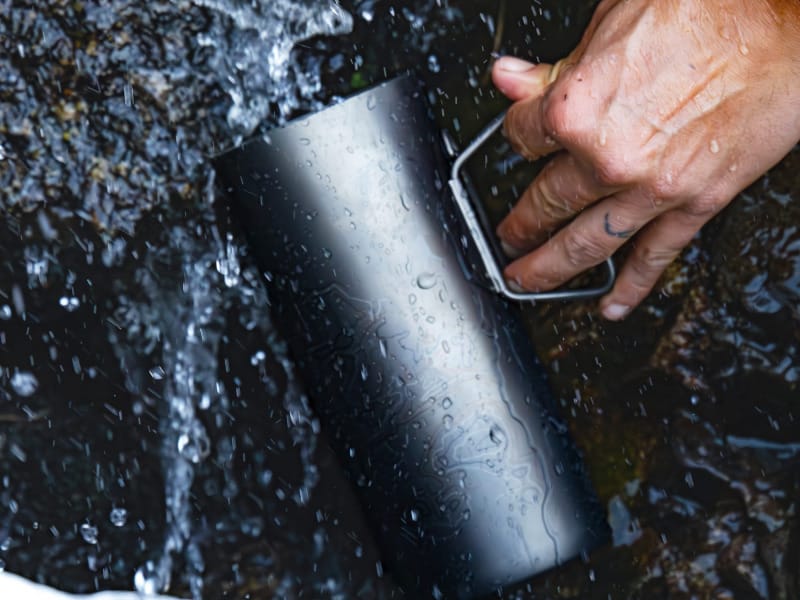 Hand submerging the GRAYL GeoPress Ti water bottle into a rushing stream to collect untreated water.