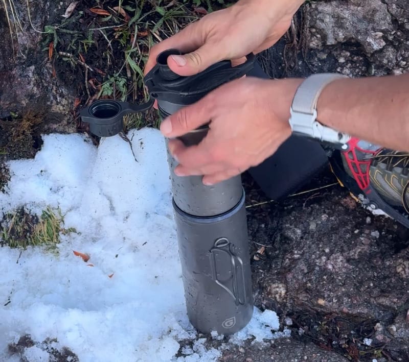 Hands pressing the GRAYL GeoPress Ti filter bottle into snow to purify water during a cold-weather hike.