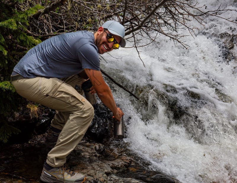 Backpacker smiling while using the GRAYL GeoPress Ti bottle to collect water from a waterfall on trail.