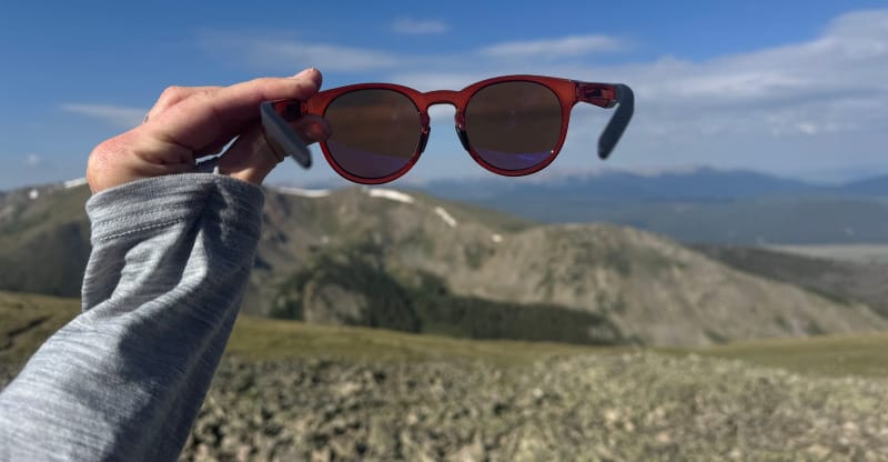 Hand holding FKT High Lonesome sunglasses against a mountain landscape on a sunny day.