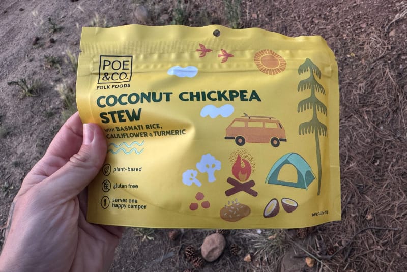  Hand holding a Poe & Co Coconut Chickpea Stew pouch outdoors on the ground with pine needles and dirt in the background.