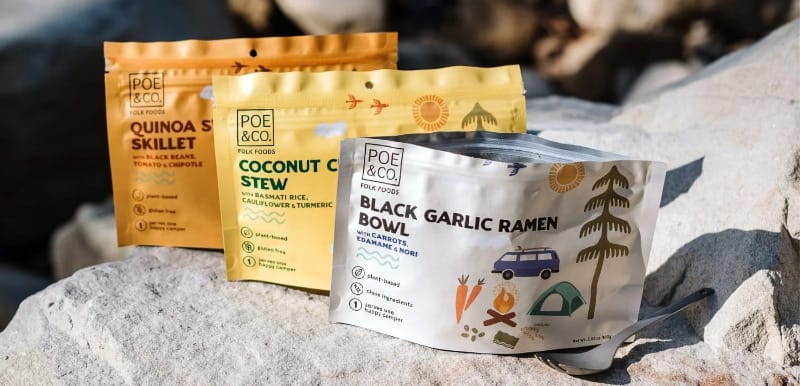 Brand image of Poe & Co’s three dehydrated backpacking meals—Quinoa Sweet Potato Skillet, Coconut Chickpea Stew, and Black Garlic Ramen—on a rock in outdoor light with a quote about the company’s chef-driven mission.