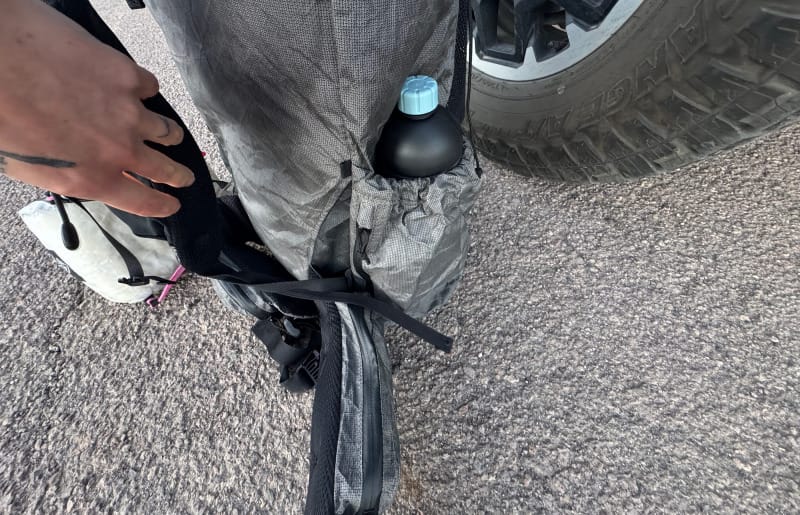  Close-up of the Kakwa 55’s side pocket holding a black titanium bottle, next to a hiker’s hand adjusting the shoulder strap.