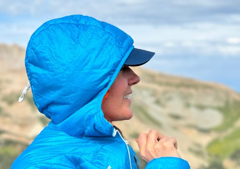 Side profile of a hiker wearing the Mammut Eigerjoch Light IN Hooded Jacket with the hood up, standing against a backdrop of high peaks.