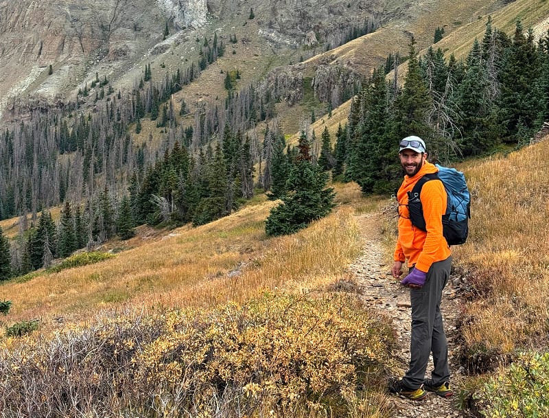 Hiker in blaze orange jacket standing on a high alpine trail, staying visible against rugged mountain terrain during hunting season.