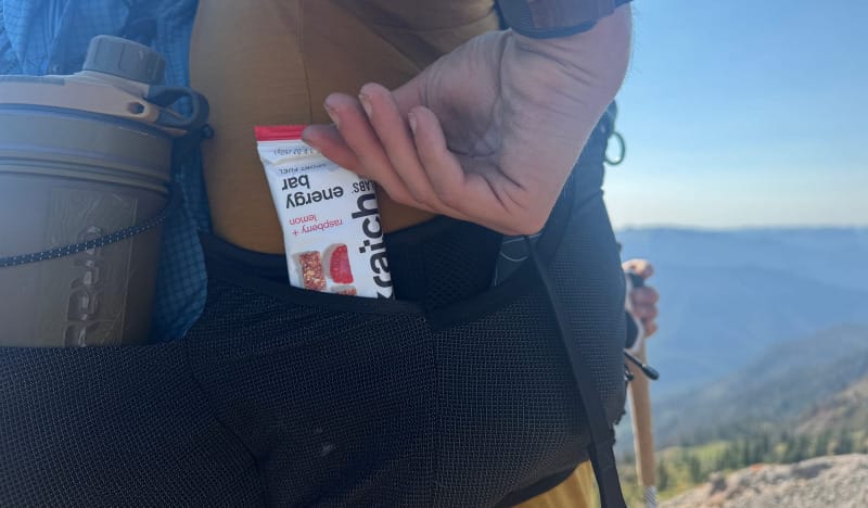 Hiker reaching into hip belt pocket to grab a Skratch Labs Raspberry + Lemon Energy Bar during a mountain trek.