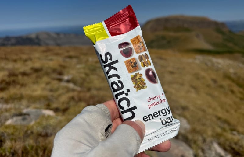 Hiker holding a Skratch Labs Cherry + Pistachio Energy Bar package with alpine ridges in the background.